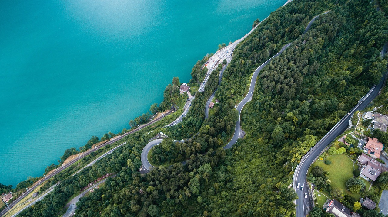 Vista aérea de uma montanha com estradas sinuosas cercadas por inúmeros pinheiros. Ao fundo, há uma lagoa cristalina que reflete o céu azul, criando uma cena natural deslumbrante.